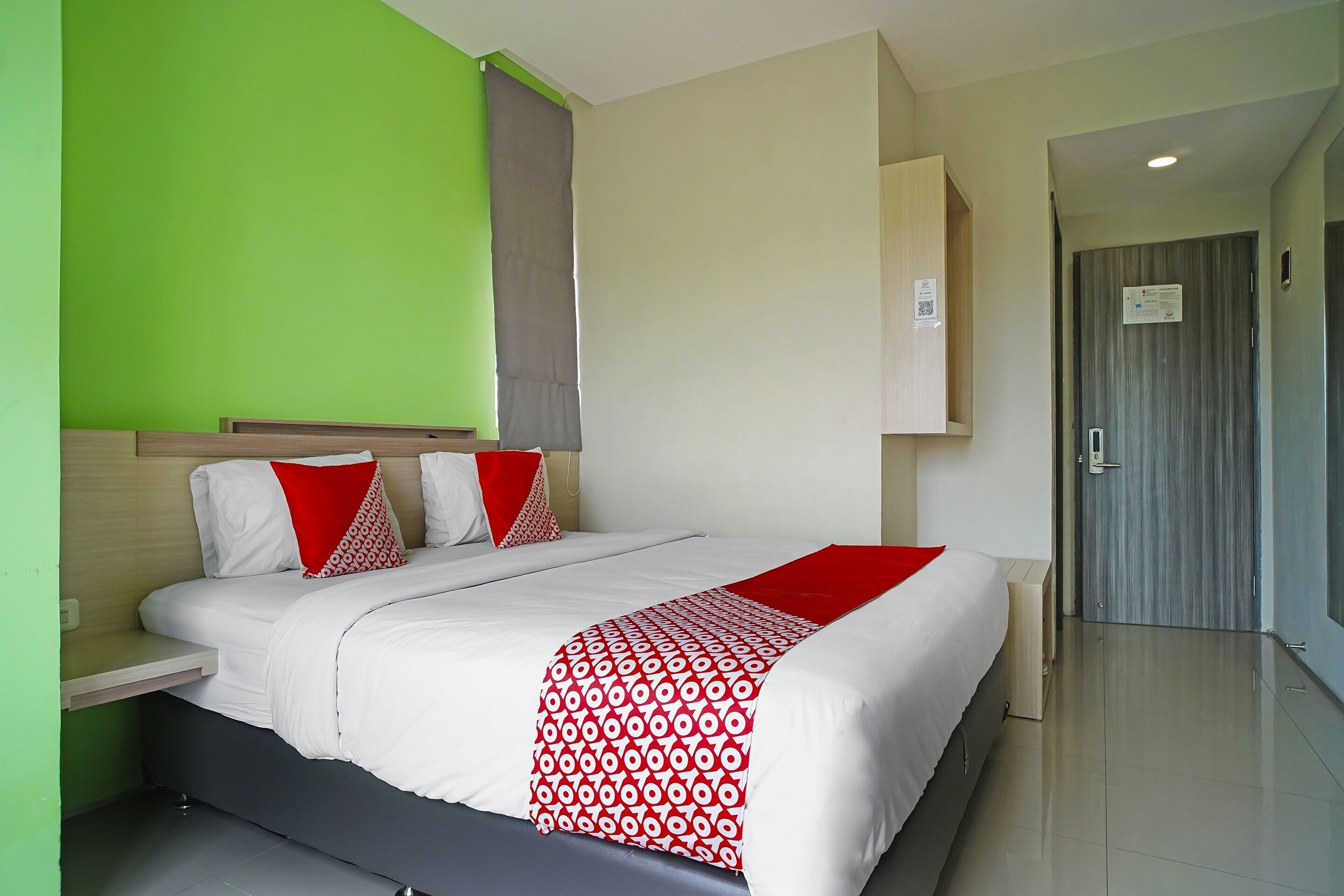 A hotel room with a bed, a nightstand, a red and white patterned throw blanket, a grey curtain, and a door with a sign.
