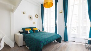 Un dormitorio con una cama, una silla blanca, una ventana con cortinas e iluminación en el techo.