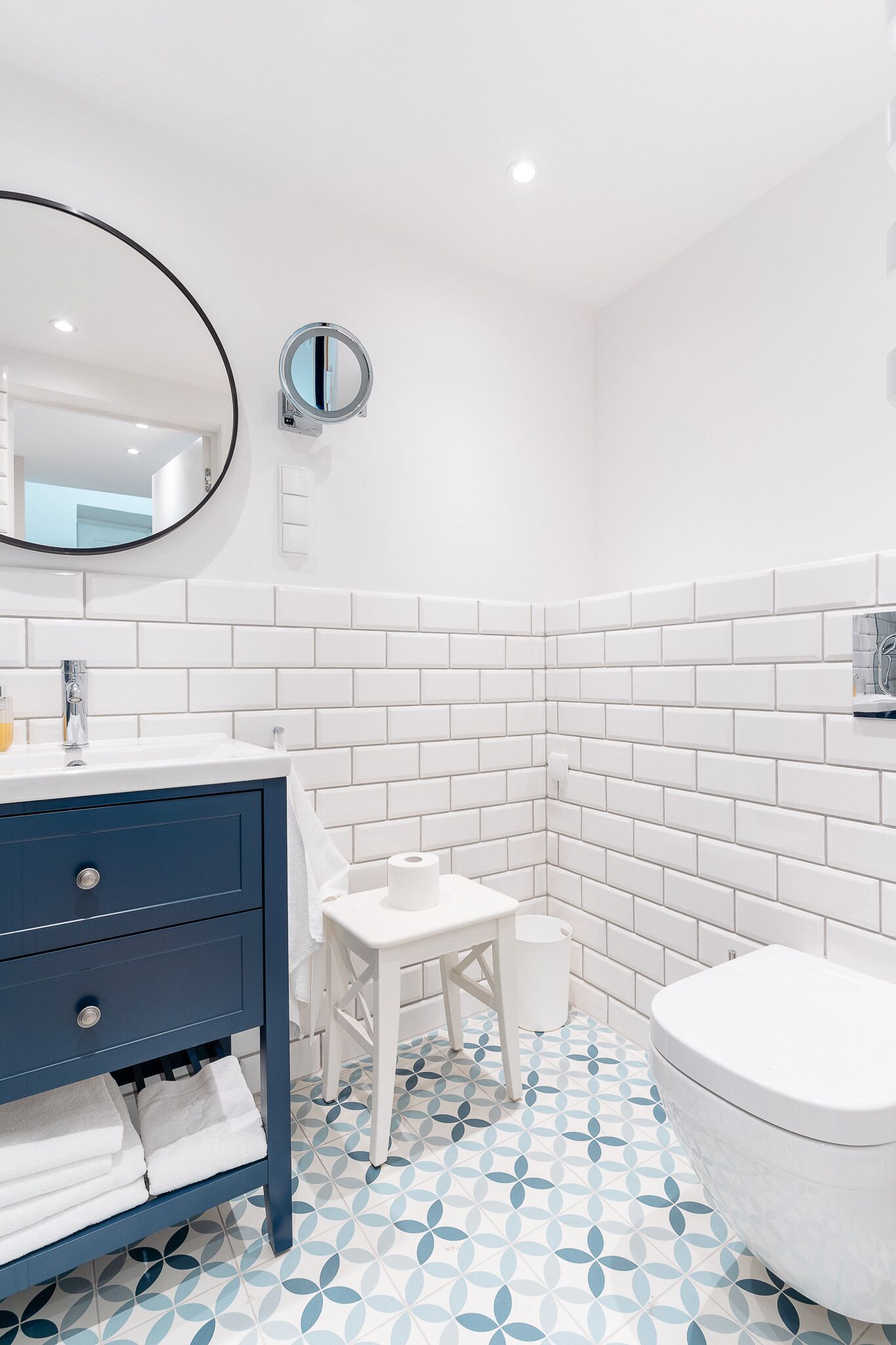 A bathroom with white tiled walls, a blue vanity, a round mirror, a toilet, and a patterned floor.