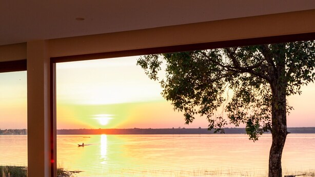 Una habitación con un ventanal grande que da a un atardecer sobre el agua, un banco de madera y un árbol afuera.