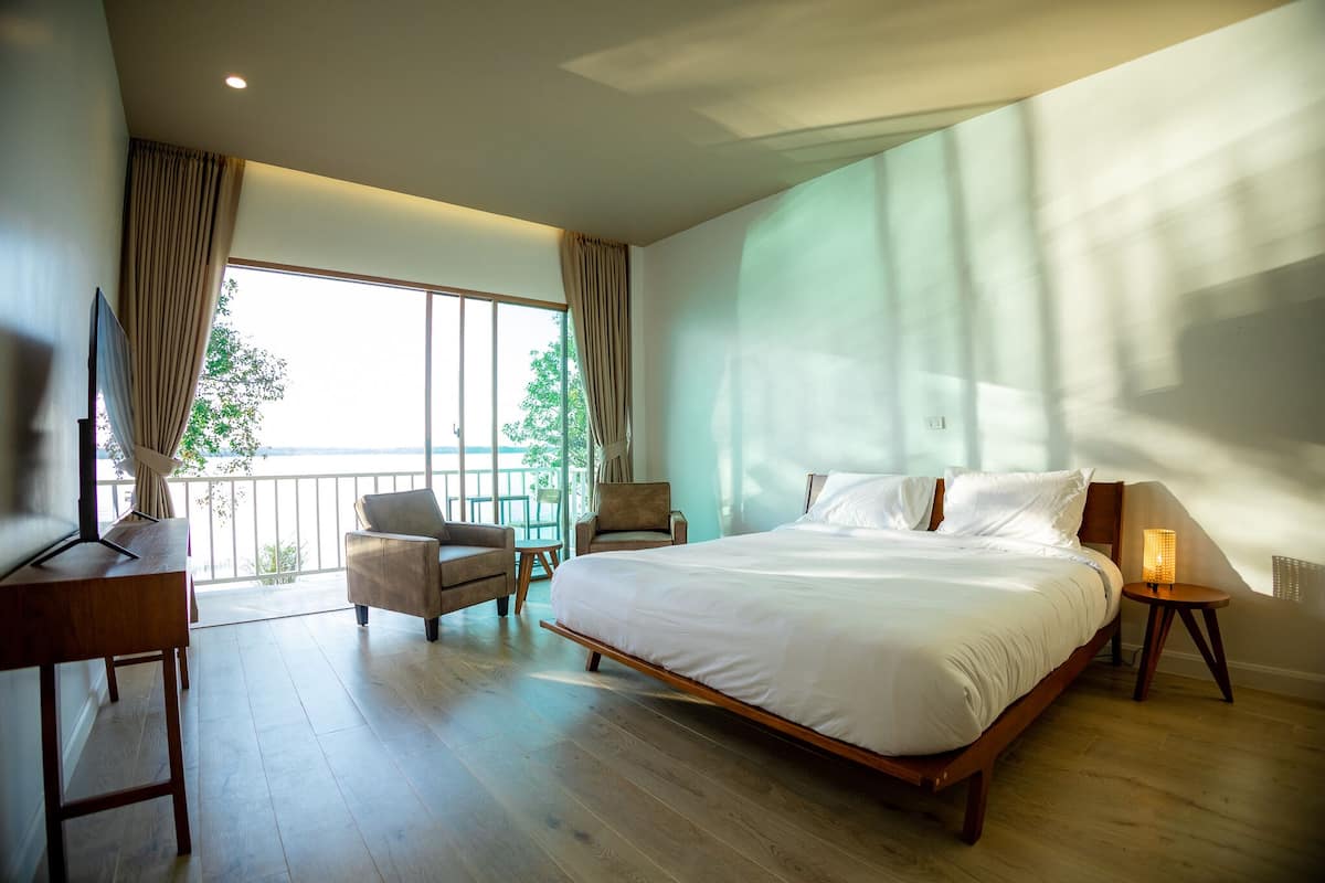 Ein Schlafzimmer mit einem großen Bett, einem Schreibtisch, zwei Stühlen, einer Stehlampe und Blick ins Freie durch ein großes Fenster.