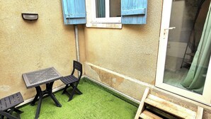 Un petit patio extérieur avec du gazon synthétique, une table et deux chaises, une fenêtre avec des volets bleus et un banc en bois.