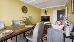 Un espace repas comprenant une table en bois, des chaises à revêtement gris, un grand téléviseur à écran plat et des œuvres d’art décoratives accrochées au mur.