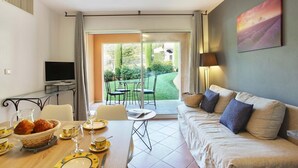 Ein modernes Wohnzimmer mit einer Couch, einem Esstisch gedeckt mit Tellern und Gläsern, einem Fernseher und Blick auf einen Garten durch eine Schiebetür.