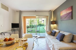 Ein modernes Wohnzimmer mit einer Couch, einem Esstisch gedeckt mit Tellern und Gläsern, einem Fernseher und Blick auf einen Garten durch eine Schiebetür.