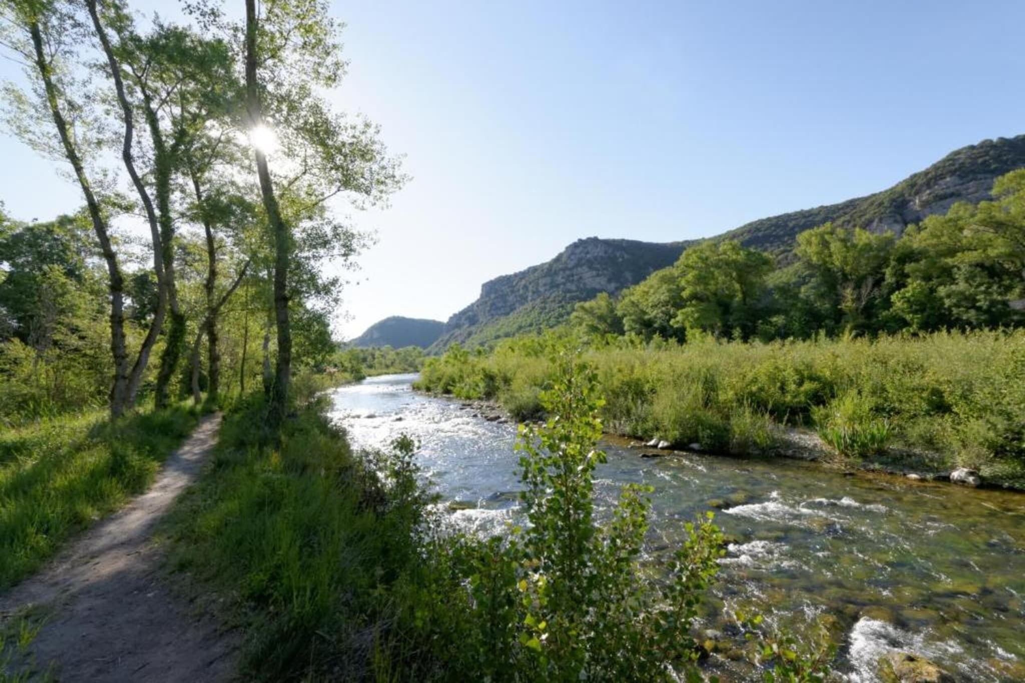 Ein Fluss fließt durch ein Tal, begleitet von einem Pfad, umgeben von Grün und Hügeln.