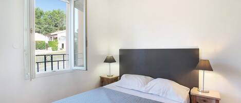 A bedroom with a bed, two bedside tables with lamps, a window with a view of greenery, and a balcony.