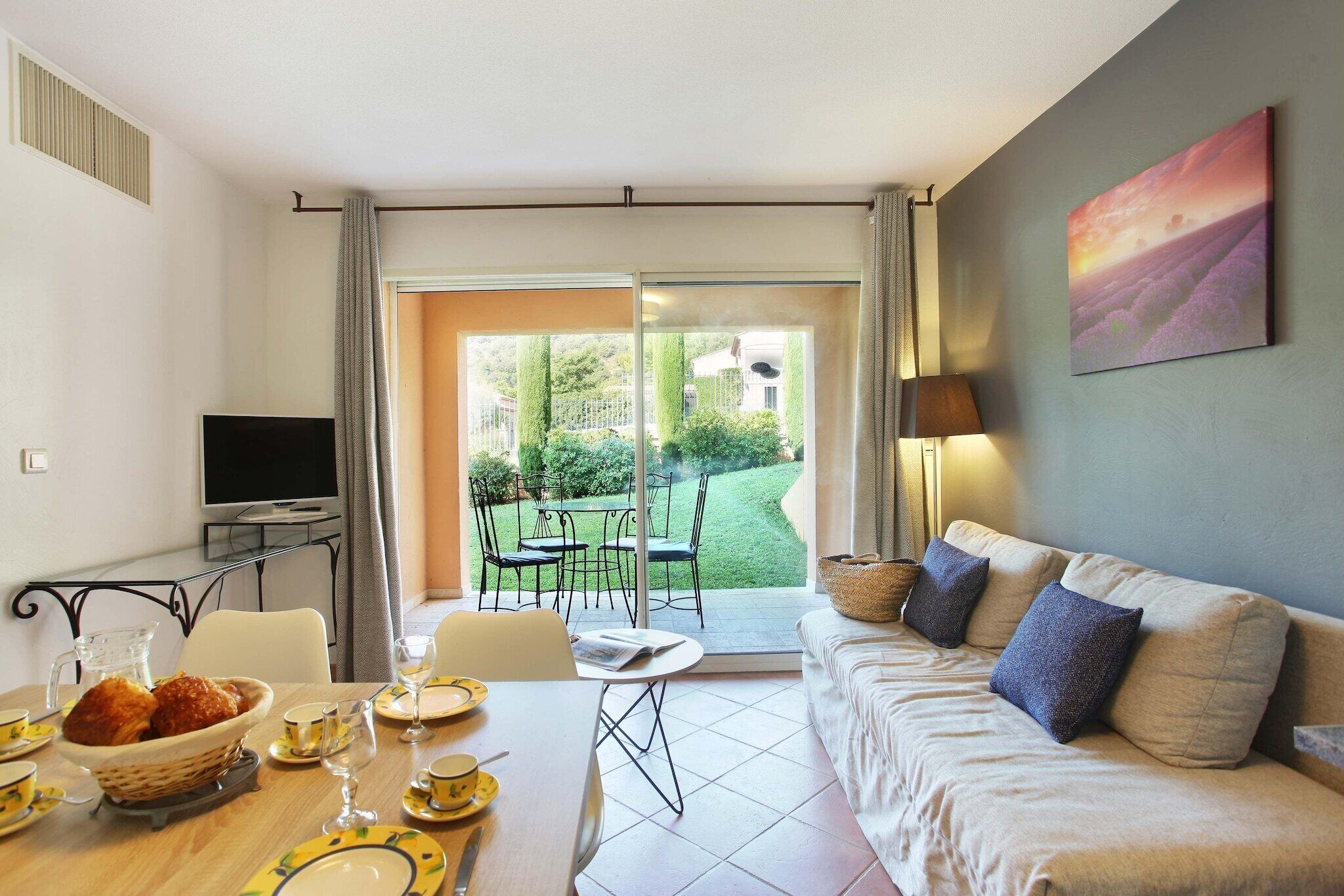 Ein modernes Wohnzimmer mit einer Couch, einem Esstisch gedeckt mit Tellern und Gläsern, einem Fernseher und Blick auf einen Garten durch eine Schiebetür.