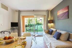 A modern living room with a sofa, a dining table set with plates and glasses, a television, and a view of a garden through a sliding glass door. - Apartment in Gréoux-Les-Bains near Lakes (Gréoux-les-Bains)