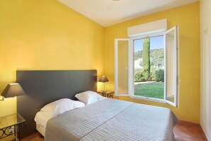 Ein Schlafzimmer mit einem Bett, zwei Nachttischlampen, einem Fenster mit geöffnetem Rollo und Blick ins Grüne.