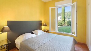 Un dormitorio con cama, dos lámparas de noche, una ventana con persiana abierta y vista a zonas verdes.