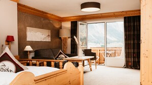 Une pièce en bois avec un lit, un canapé, une table et une chaise. Il y a une fenêtre d’où l’on peut admirer la vue sur les montagnes.