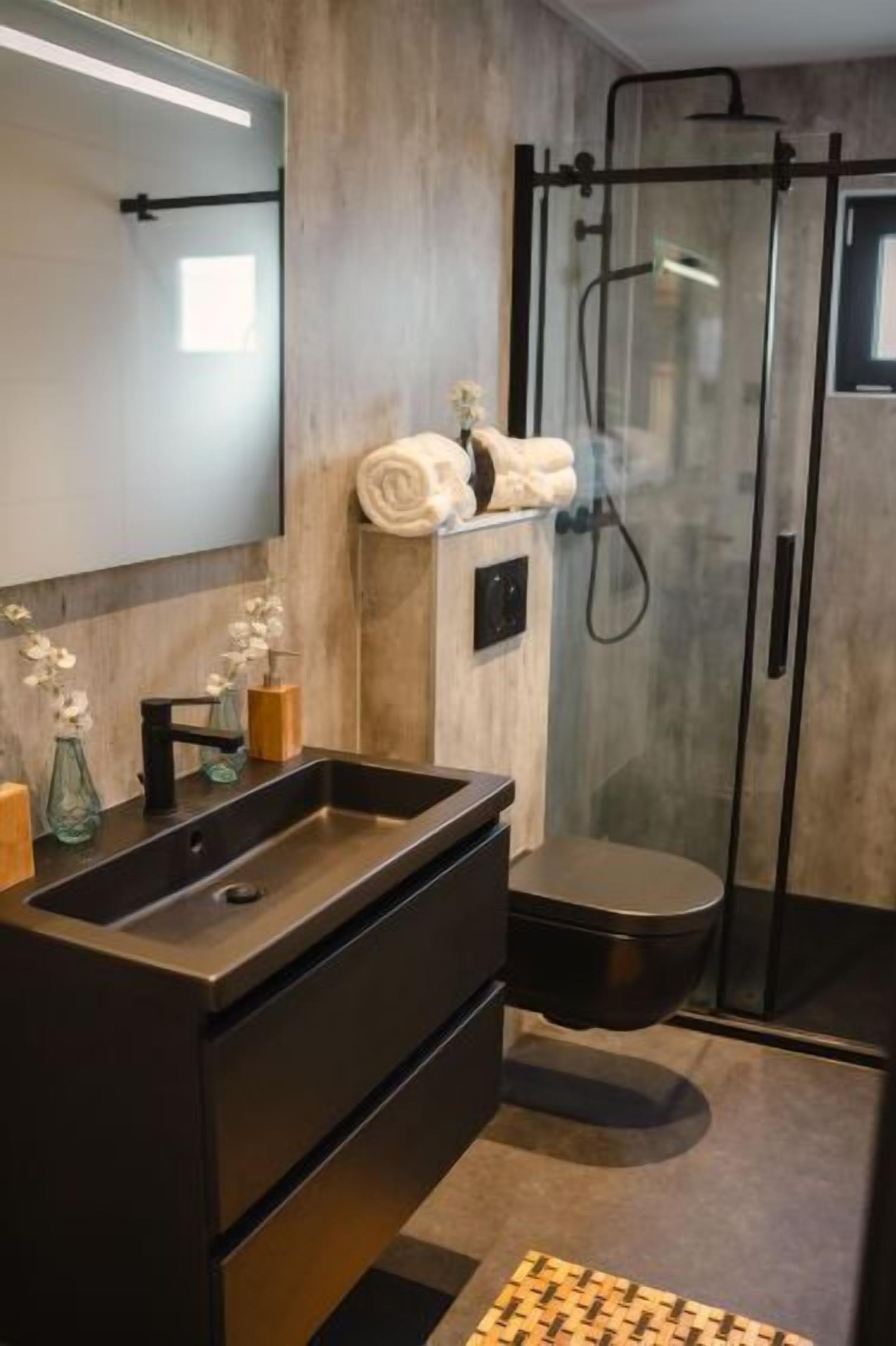 A modern bathroom with a dark sink, mirror, and toilet. There is a glass shower enclosure and a towel rack with towels.