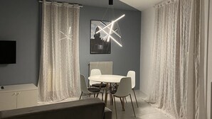 A modern living room with a dining area, featuring a contemporary light fixture, a flat-screen TV mounted on the wall, and a large window with sheer curtains. - Serendipity Apartment-Your home in Venice (Venezia)