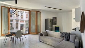 A modern living room with a sofa, dining table, and chairs. There is a kitchen area with a refrigerator and a sink. The room has large windows with a view of buildings outside. - Serendipity Apartment-Your home in Venice (Venezia)