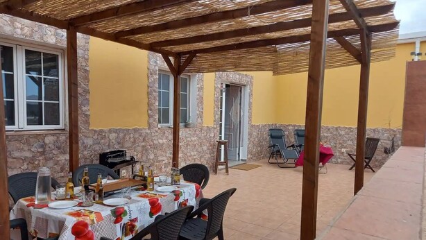 Une terrasse extérieure avec une table dressée pour les repas, des murs en pierre et un espace couvert avec des chaises.
