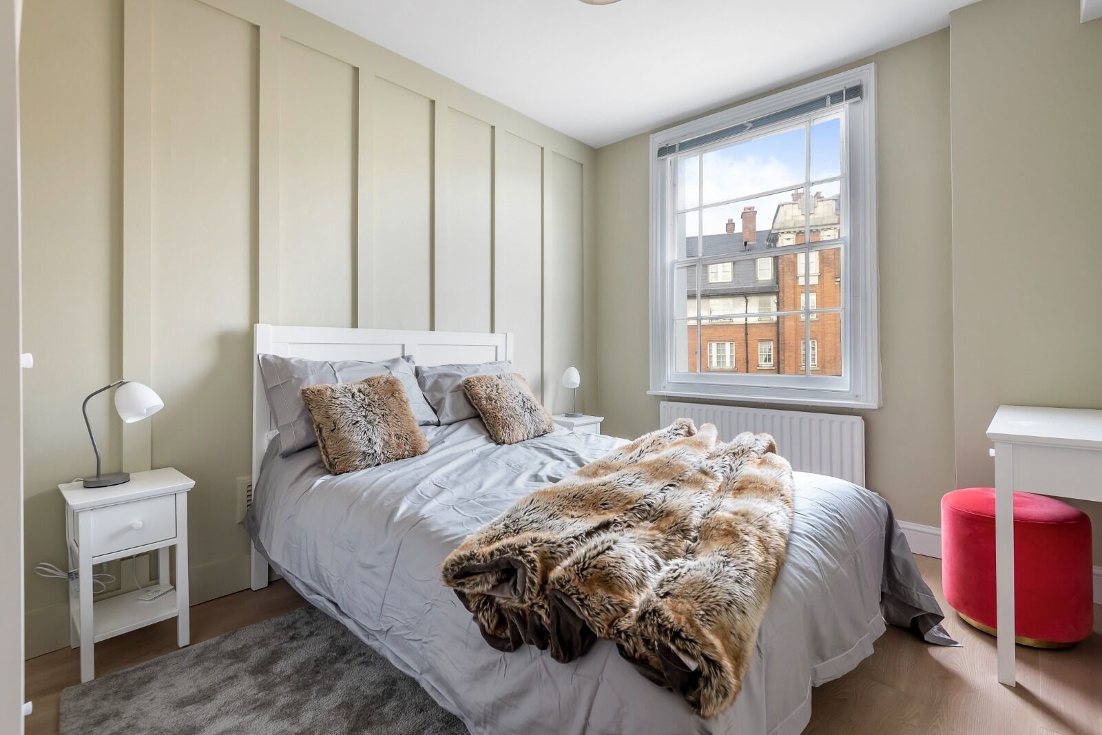 Une chambre à coucher comprenant un lit, une table de chevet, un pouf rouge, une fenêtre donnant sur des immeubles et un radiateur.