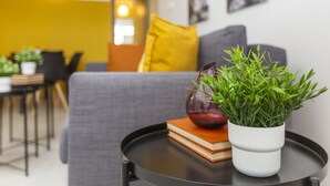Un salon moderne avec un canapé gris, un coussin jaune, un vase blanc contenant des plantes vertes et une pile de livres sur une table d’appoint noire.