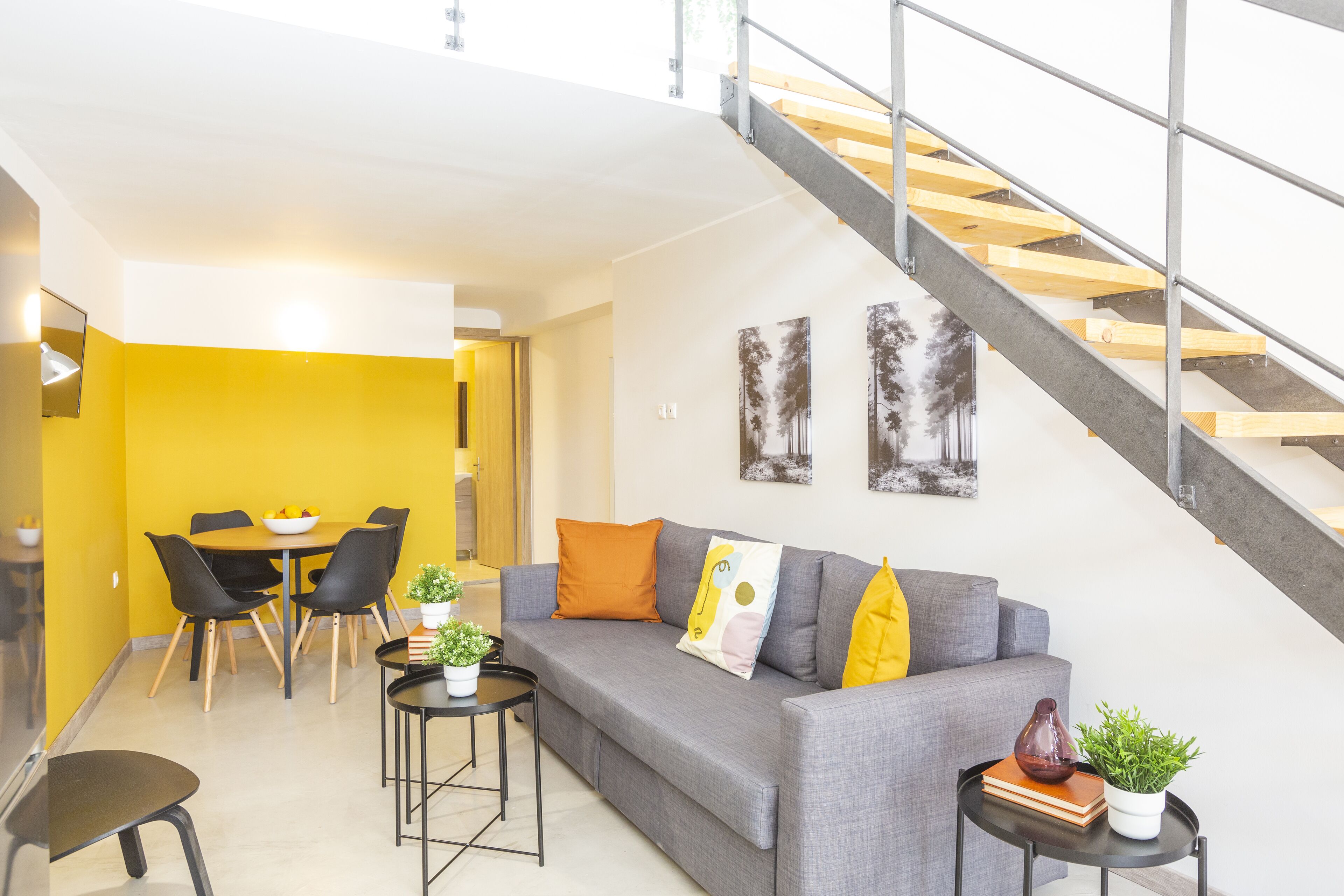 A modern living room with a grey sofa, black chairs, a dining table with chairs, a small round table with a plant, and a staircase with a wooden railing.