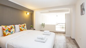 Une chambre d’hôtel moderne avec un grand lit, des oreillers colorés, une petite table de chevet agrémentée d’une plante, et une vue sur l’extérieur.