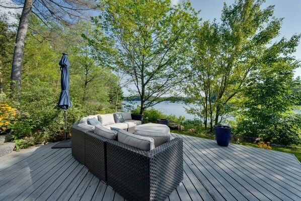 A deck with a wicker sofa and a patio umbrella, surrounded by trees and a view of a lake.