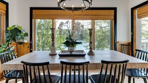 A dining area with a large table, six chairs, a chandelier, and large windows with views of trees.