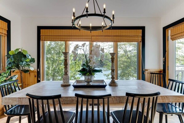 A dining area with a large table, six chairs, a chandelier, and large windows with views of trees. - La Belle Vue - Beautiful Sunsets on Long Lake (Torrance)