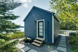 A small, dark blue cabin with a white door and windows, surrounded by a gravel path and greenery, overlooking a body of water. - La Belle Vue - Beautiful Sunsets on Long Lake (Torrance)
