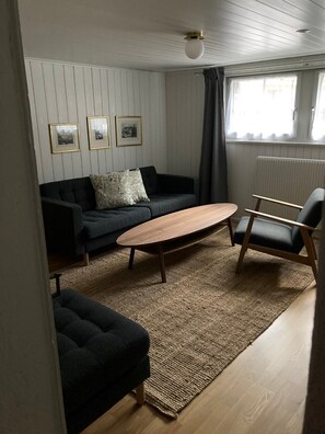 Una sala de estar con un sofá gris oscuro, una mesa de centro de madera y un sillón negro. Hay tres cuadros enmarcados en la pared y una ventana con cortinas.