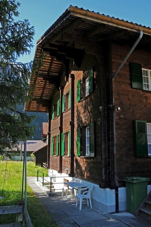 Un edificio de dos pisos de ladrillo con contraventanas verdes, un espacio de estar al aire libre cubierto con sillas blancas y un bote de basura visible.
