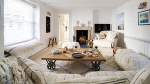 A cozy living room with a fireplace, a sofa set, a coffee table with books and wine glasses, and a television mounted on the wall. - This charming 18th Century cottage is centrally located in the heart of St Teath (St Teath)