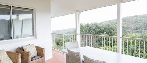 A balcony with wicker furniture and a white table, overlooking a lush green landscape.