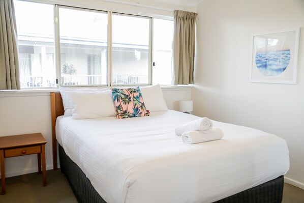 Una cama bien tendida con sábanas blancas y una almohada tropical, una mesita de noche y una ventana con cortinas.