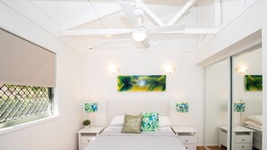 A bedroom with a bed, white nightstands, a ceiling fan, artwork on the walls, and a window with a metal screen. - Straddie Breeze - A gorgeous fully renovated beachside apartment (Point Lookout)