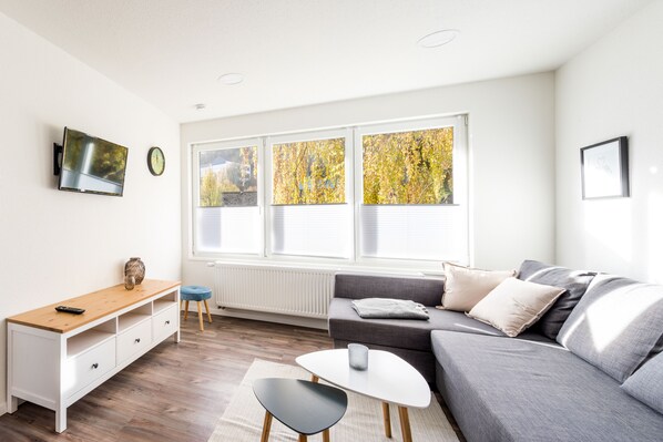 Un salon moderne avec un canapé gris, une table basse en bois, un meuble TV blanc et une fenêtre donnant sur des arbres aux couleurs automnales.