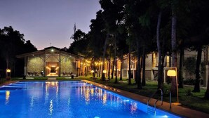 Une piscine à l’eau d’un bleu limpide, entourée d’arbres et bordée d’un bâtiment à l’éclairage chaleureux.
