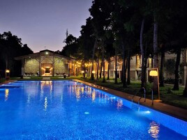 Uma piscina com superfície de água azul cristalina, rodeada por árvores e um edifício com iluminação acolhedora.