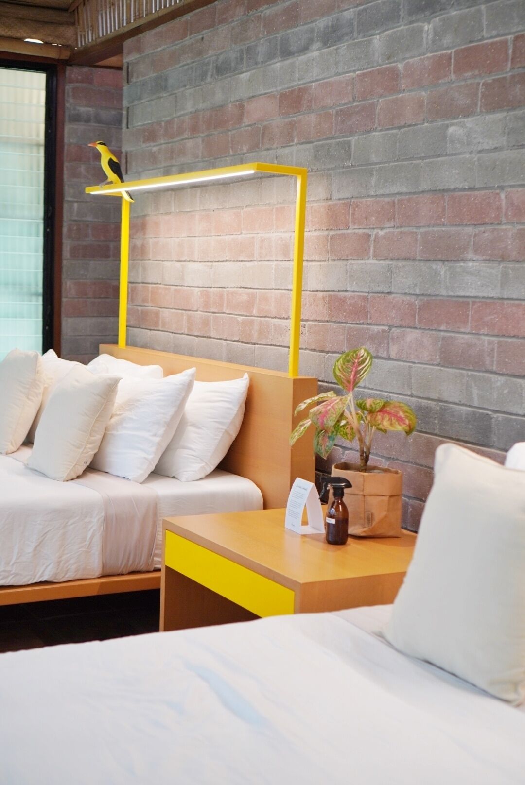 Une chambre d’hôtel avec un mur en briques, un lit en bois avec une literie blanche, une petite table de chevet agrémentée d’une plante et une tête de lit jaune.