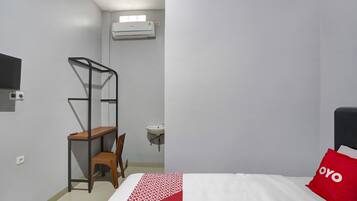 A bedroom with a bed, a red pillow, a desk with a chair, a hanging air conditioner, and a wall-mounted sink.
