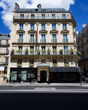 Un edificio de varios pisos con estilo arquitectónico clásico, que presenta una entrada central con toldo negro, múltiples balcones con barandillas de hierro forjado y una fila de ventanas con contraventanas azules.