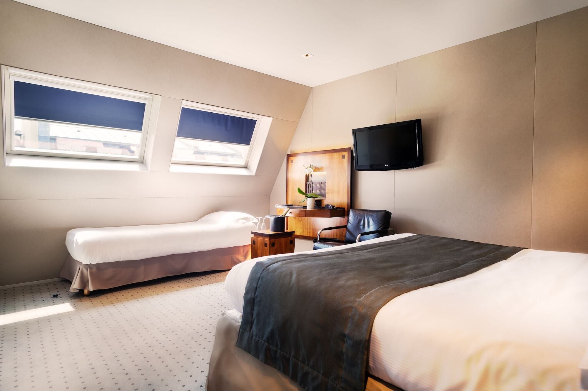 A hotel room with two beds, a desk with a chair, a TV mounted on the wall, and a window with a view of buildings.