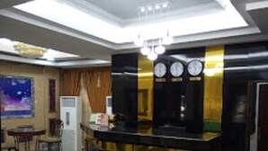 Un vestíbulo de hotel con mostrador de recepción, un reloj, una pequeña mesa con sillas y un cartel en la pared.