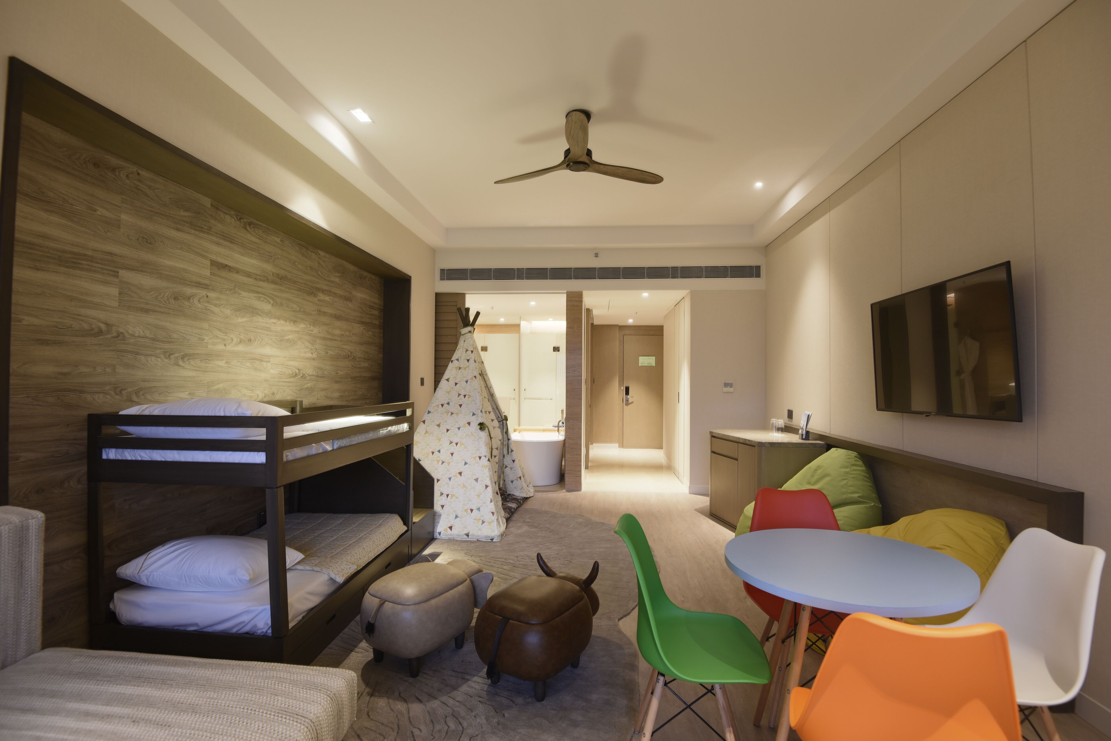 A modern hotel room with bunk beds, a small table, colorful chairs, a ceiling fan, and a TV.
