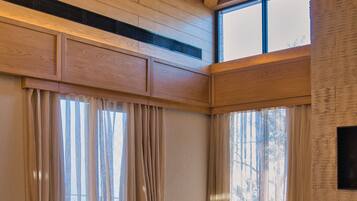 A room with wooden ceiling beams, large windows with sheer curtains, a woven chair, a bean bag, and a round wooden table.
