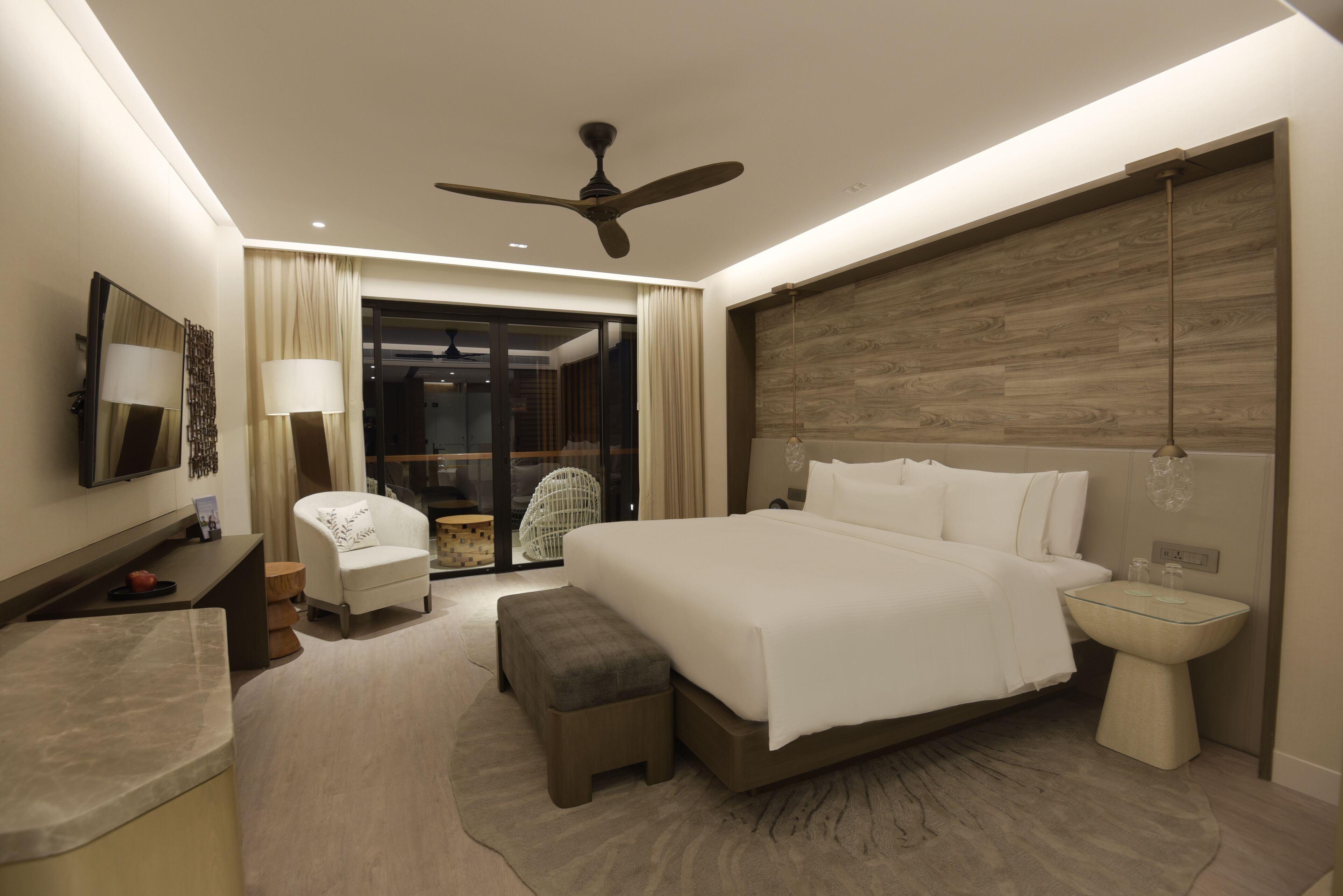 A modern hotel room with a large bed, a ceiling fan, a flat-screen TV, a desk with a lamp, and a view of the outside through a glass door.