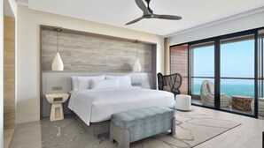 Un dormitorio moderno con una cama grande, ventilador de techo, vista al océano y balcón con mobiliario de mimbre.
