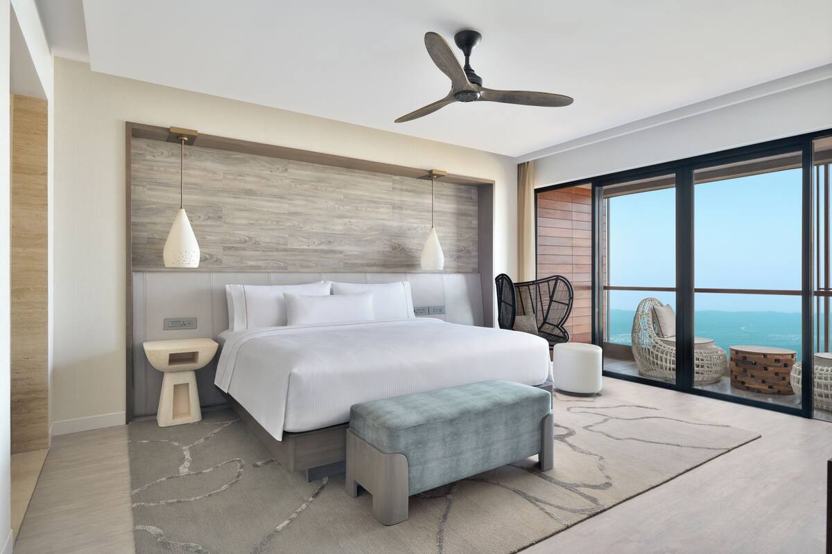 A modern bedroom with a large bed, a ceiling fan, a view of the ocean, and a balcony with wicker furniture.