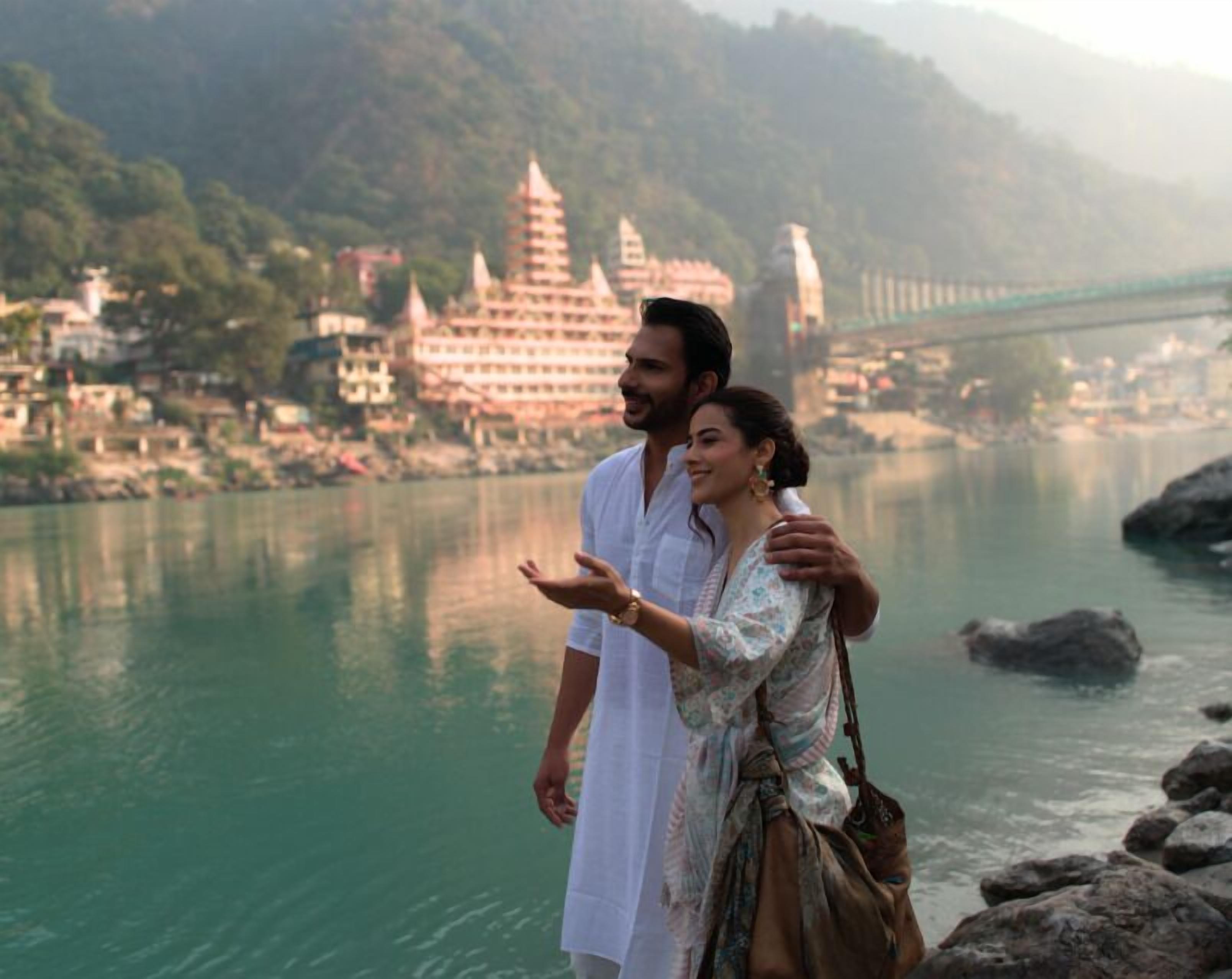 A couple standing by a river with a bridge and temples in the background.