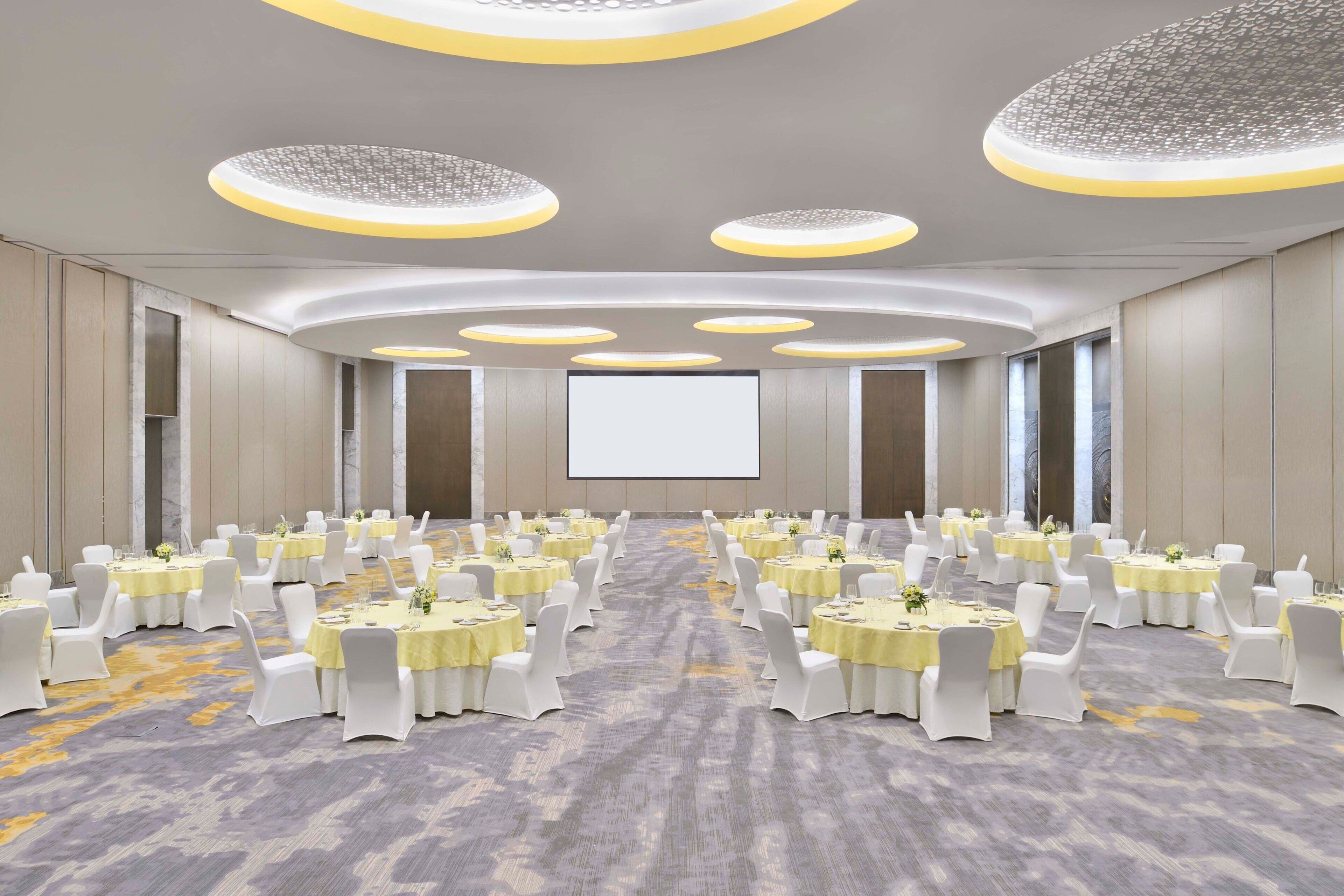 Un salón de conferencias con mesas redondas, sillas blancas y una pantalla grande al frente.