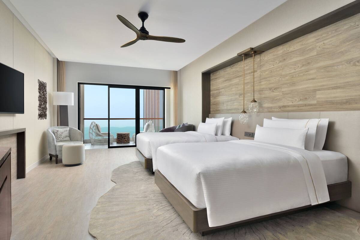 A modern hotel room with two beds, a ceiling fan, a large window with a view, and a flat-screen TV.